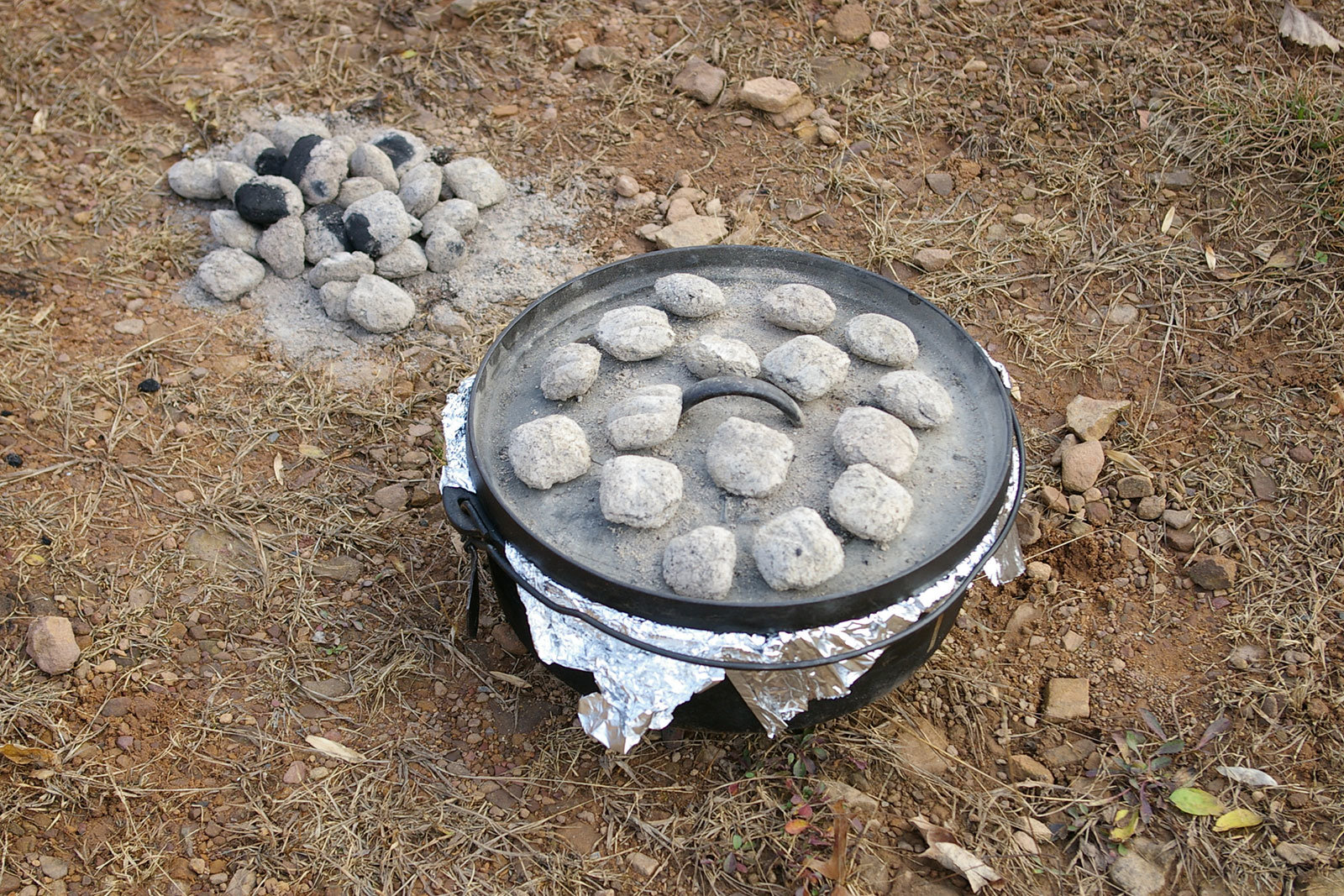 A Dutch-oven covered with tin-foil, a lid and charcoals, and other charcoals next to it.
