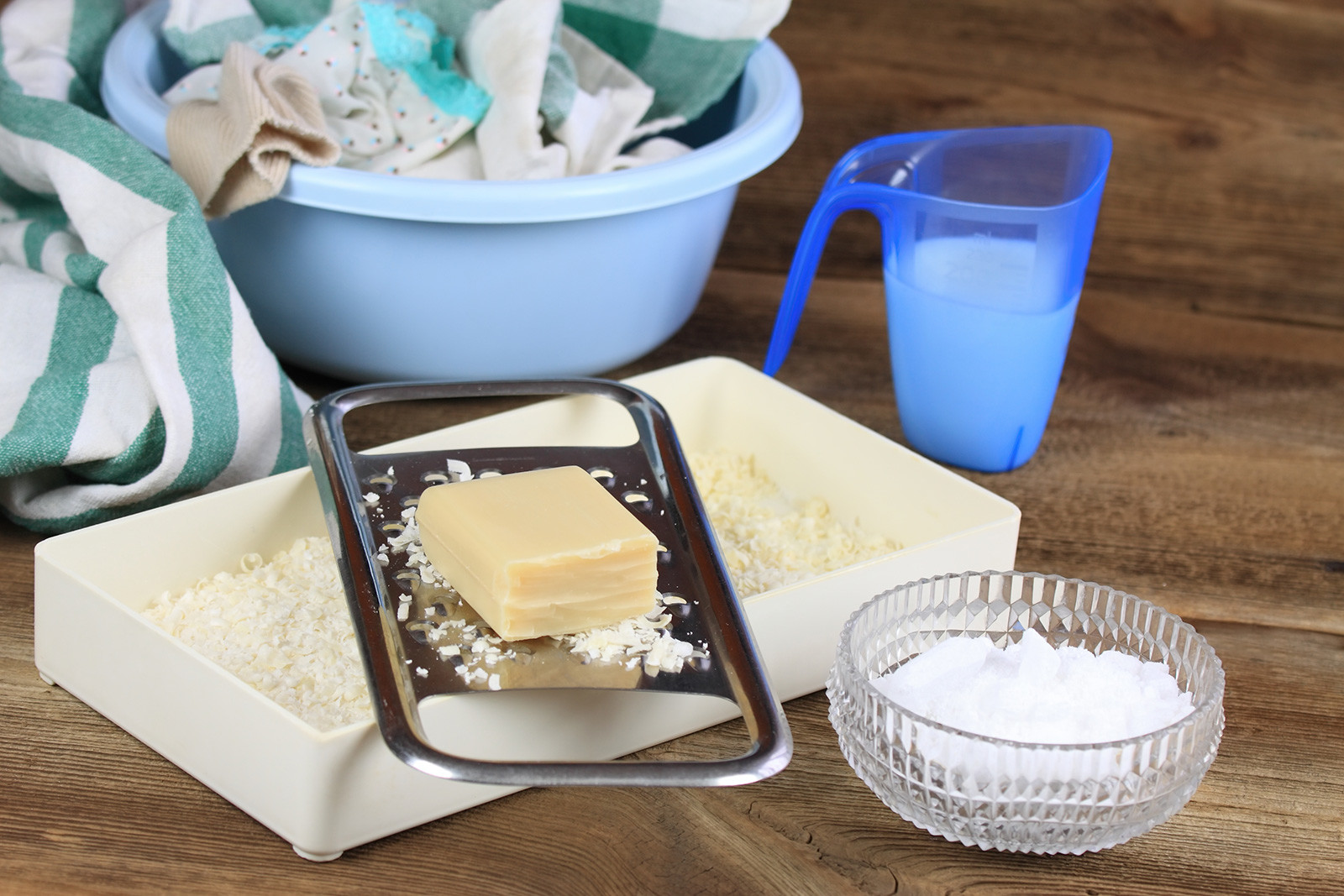 Soap and grated soap, crystalline-sodium in a bowl, and some clothes/cloths in a shallow bucket.