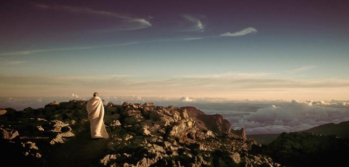 A person meditating, standing on a mountain peak, in front of the horizon.